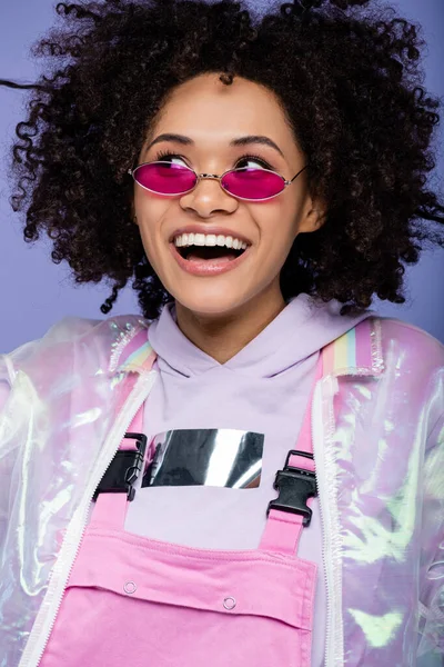 Жизнерадостная африканская американка в солнцезащитных очках, смотрящая вдаль изолированной на фиолетовый — стоковое фото