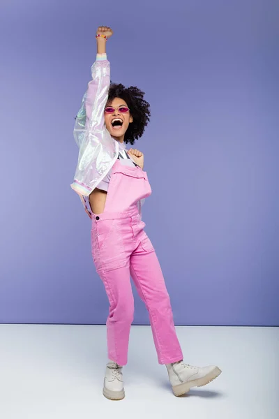 Full length of excited african american woman in stylish overalls on purple — Stock Photo