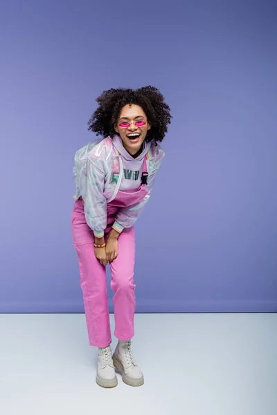 Full length of cheerful african american woman in stylish overalls laughing on purple — Stock Photo