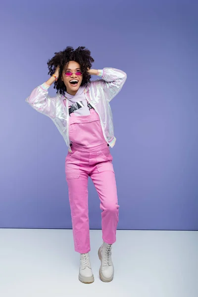 Full length of cheerful african american woman in stylish overalls posing and adjusting curly hair on purple — Stock Photo