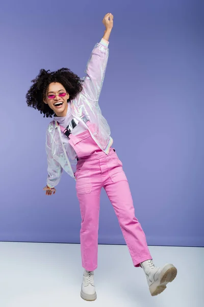 Pleine longueur de femme afro-américaine excitée en salopettes élégantes et lunettes de soleil posant sur violet — Photo de stock