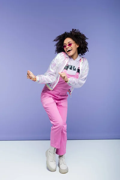 Comprimento total de mulher americana africana positiva em macacões elegantes posando em roxo — Fotografia de Stock