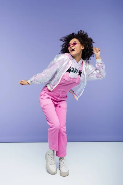 Full length of positive african american woman in trendy pink overalls posing on purple — Stock Photo