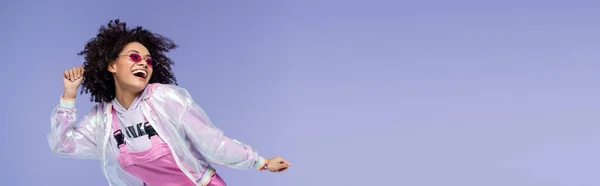 Весёлая африканская американка в розовых солнцезащитных очках жестикулирует изолированным на фиолетовом, баннере — стоковое фото