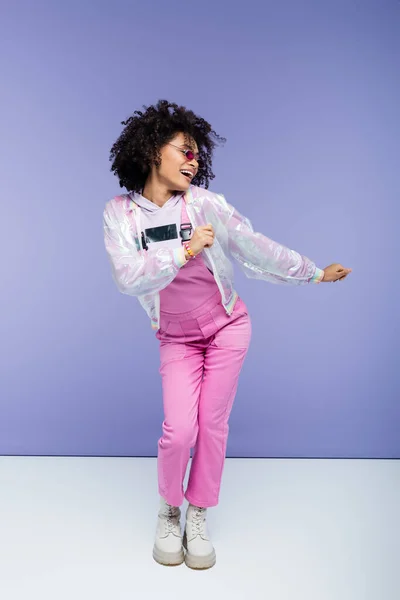 Full length of smiling african american woman in stylish overalls posing on purple — Stock Photo