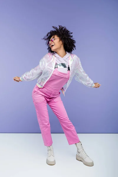 Full length of pleased african american woman in stylish overalls posing on purple — Stock Photo