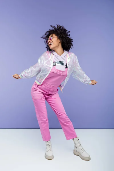 Longitud completa de la mujer afroamericana complacida en overoles elegantes posando en púrpura - foto de stock