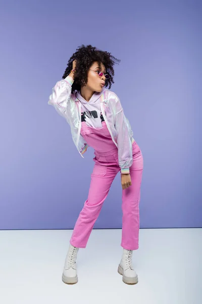 Full length of surprised african american woman in stylish overalls posing on purple — Stock Photo