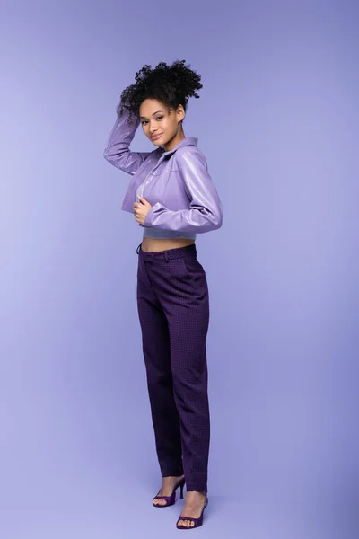 Longitud completa de la alegre mujer afroamericana en chaqueta de cuero violeta posando en púrpura - foto de stock