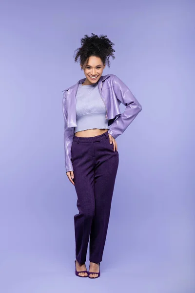 Full length of cheerful african american woman in violet leather jacket and trousers posing with hand on hip on purple — Stock Photo