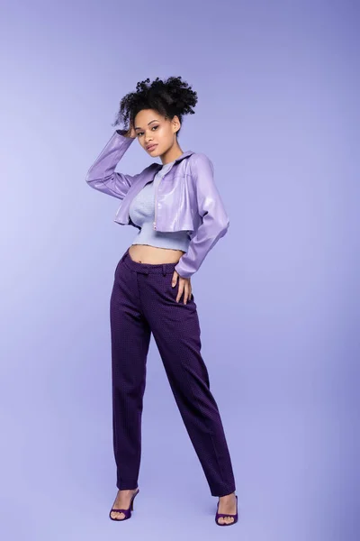 Full length of curly african american woman in violet leather jacket and trousers posing with hand on hip on purple — Stock Photo