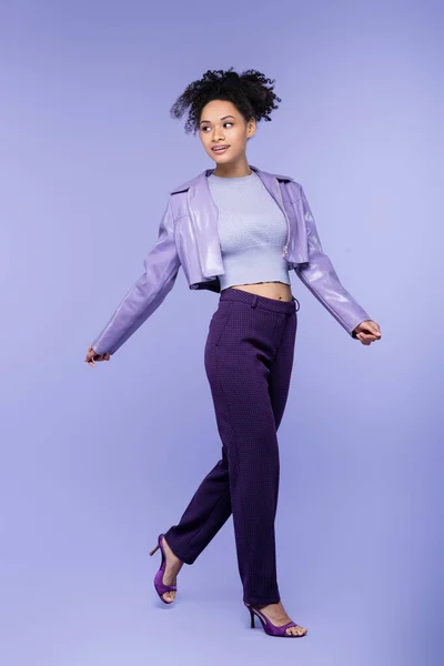 Full length of cheerful african american woman in violet leather jacket and trousers walking on purple — Stock Photo