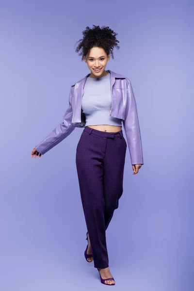 Pleine longueur de femme afro-américaine heureuse en veste et pantalon en cuir violet marchant sur violet — Photo de stock