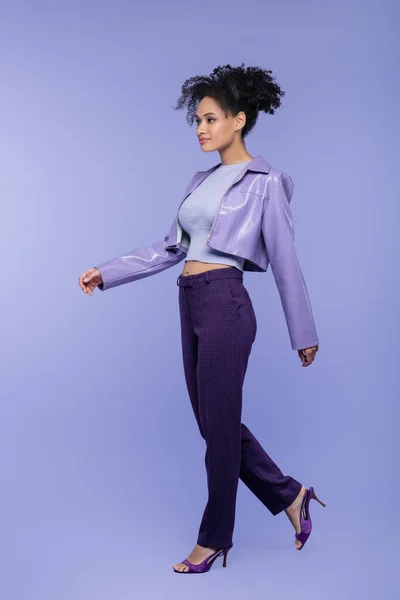 Full length of curly african american woman in violet leather jacket and trousers walking on purple — Stock Photo