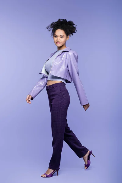 Full length of stylish african american woman in violet leather jacket and trousers walking on purple — Stock Photo
