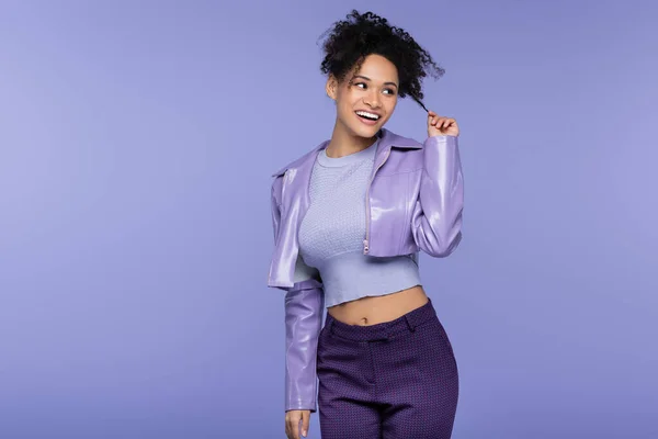 Mulher americana africana alegre em jaqueta de couro violeta ajustando o cabelo isolado em roxo — Fotografia de Stock