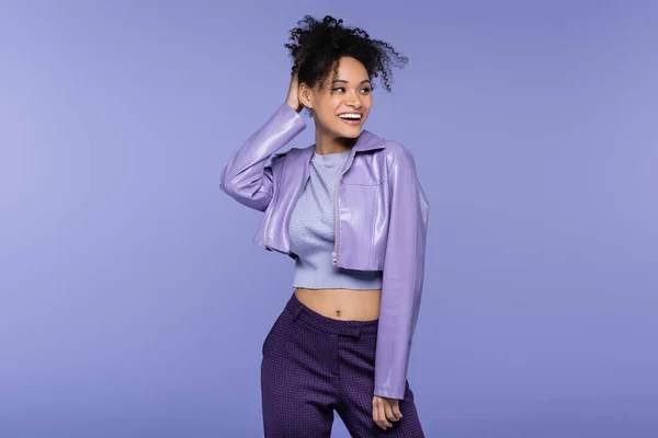 Trendy african american woman in leather jacket smiling and looking away isolated on purple — Stock Photo