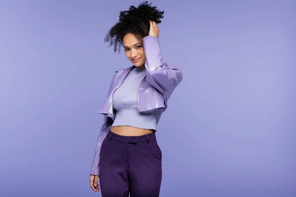 Trendy african american woman in leather jacket smiling while adjusting curly hair isolated on purple — Stock Photo