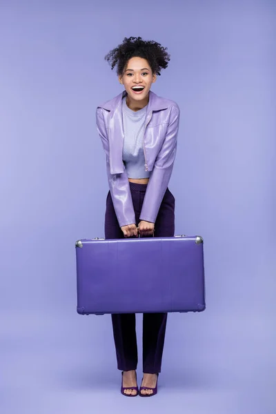 Полная длина взволнованной африканской американки, держащей чемодан на фиолетовом — стоковое фото