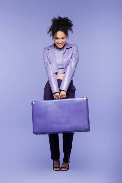 Full length of cheerful african american woman holding luggage on purple — Stock Photo