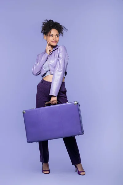 Full length of pensive young african american woman holding baggage on purple — Stock Photo