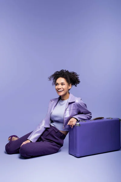 Повна довжина щасливої афроамериканської жінки, що сидить біля валізи на фіолетовому — стокове фото