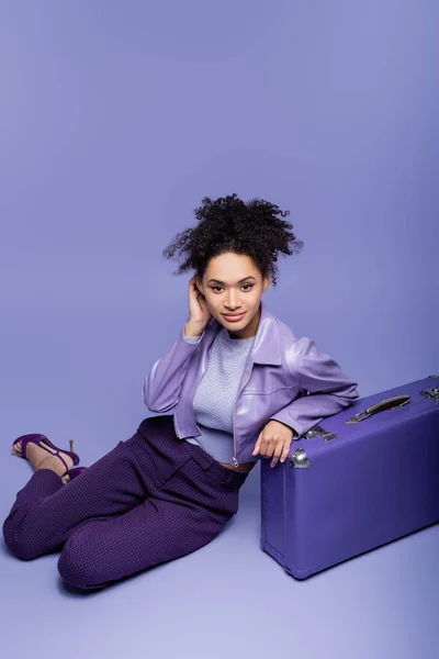Повна довжина кучерявої афроамериканської жінки, що сидить біля валізи на фіолетовому — стокове фото