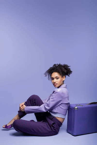 Full length of young and curly african american woman sitting near suitcase on purple — Stock Photo