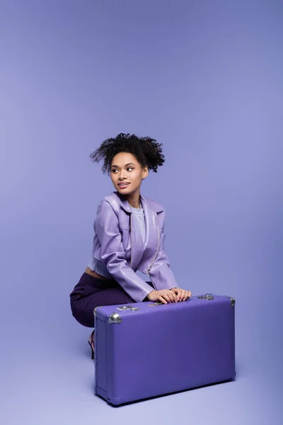 Full length of curly young african american woman sitting near baggage on purple — Stock Photo