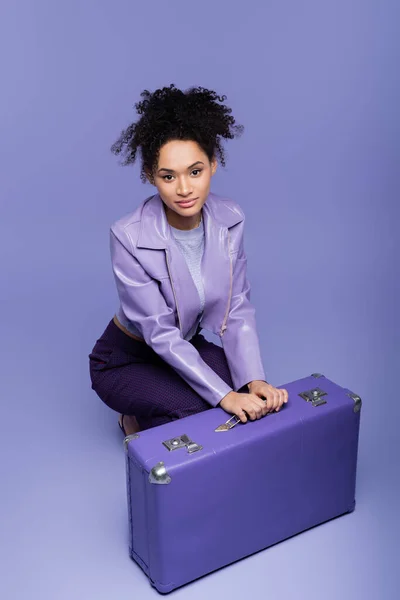 Full length of african american young woman sitting near suitcase on purple — Stock Photo