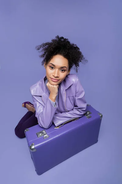 Hochwinkelige Ansicht der jungen afrikanisch-amerikanischen Frau sitzt in der Nähe von Koffer auf lila — Stockfoto