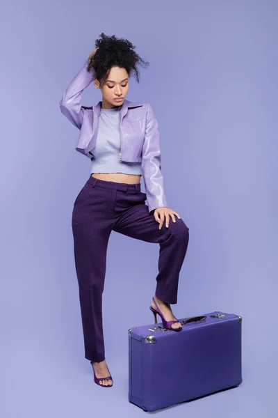 Повна довжина стильної афроамериканської жінки, що стоїть біля валізи на фіолетовому — стокове фото