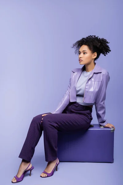 Full length of young african american woman sitting on suitcase on purple — Stock Photo