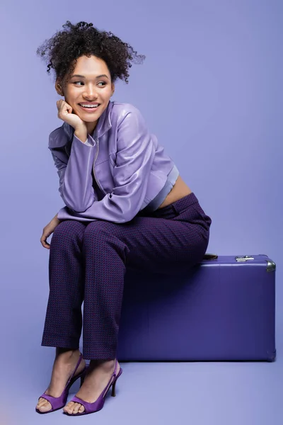 Full length of happy african american woman sitting on suitcase on purple — Stock Photo