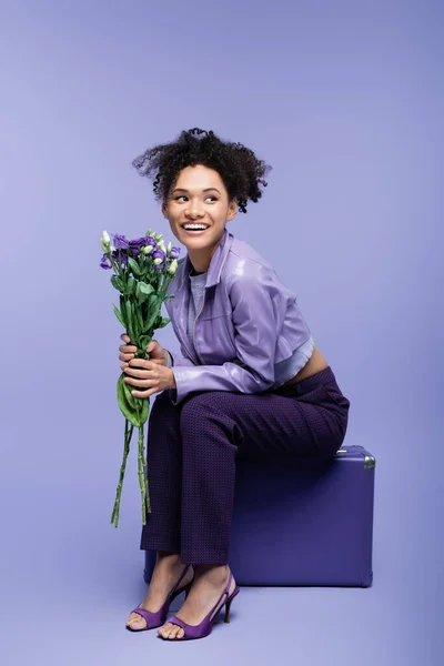 Pleine longueur de femme afro-américaine heureuse assise sur une valise et tenant des fleurs sur violet — Photo de stock