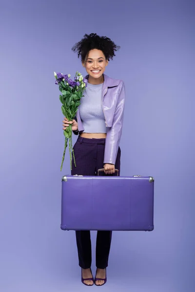 Full length of smiling african american woman holding baggage and bouquet of flowers on purple — Stock Photo