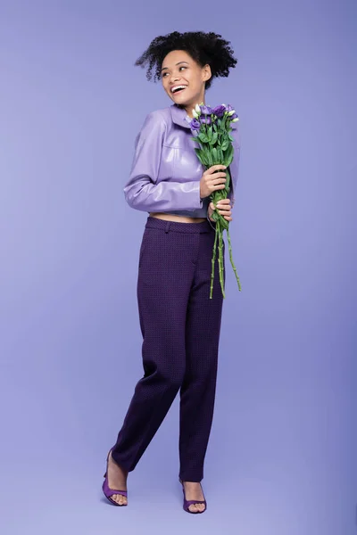 Longitud completa de la sonriente mujer afroamericana sosteniendo ramo de flores en púrpura - foto de stock