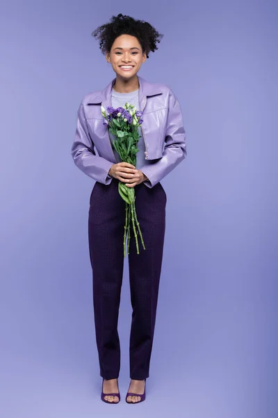 Pleine longueur de femme afro-américaine heureuse tenant bouquet de fleurs sur violet — Photo de stock