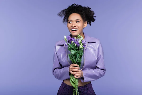 Glückliche junge afrikanisch-amerikanische Frau hält Blumenstrauß in der Hand und schaut isoliert auf lila — Stockfoto