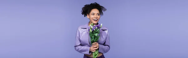 Glückliche junge afrikanisch-amerikanische Frau hält Blumenstrauß in der Hand und schaut isoliert auf violettem Banner weg — Stockfoto