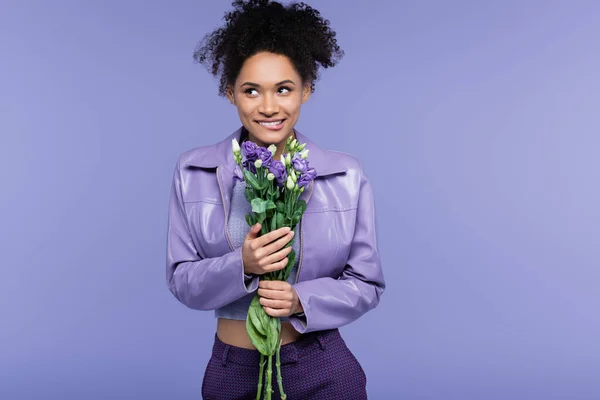 Happy young african american woman biting lip and holding bouquet of flowers isolated on purple — Stock Photo