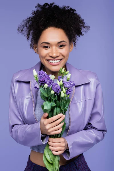 Позитивна молода афроамериканка тримає букет фіолетових квітів ізольовано на фіолетовому — стокове фото