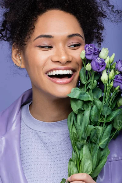 Excitada joven afroamericana mujer cerca de ramo de flores violetas aisladas en púrpura - foto de stock