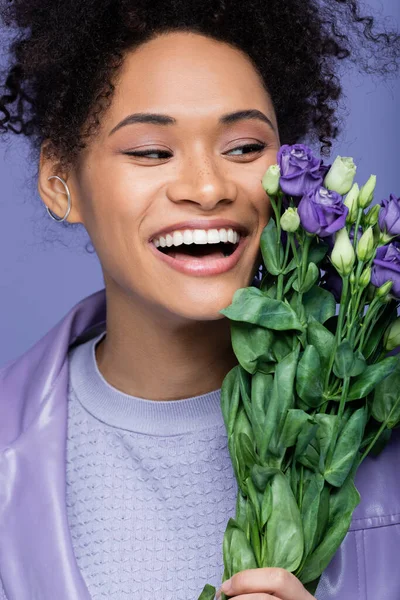 Animado jovem mulher afro-americana perto de buquê de flores violetas isoladas em roxo — Fotografia de Stock