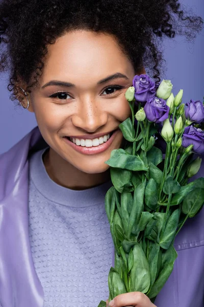 Positive and curly african american woman near bouquet of violet flowers isolated on purple — Stock Photo