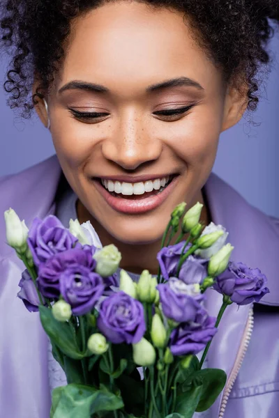 Positive jeune femme afro-américaine regardant flous fleurs violettes isolées sur violet — Photo de stock