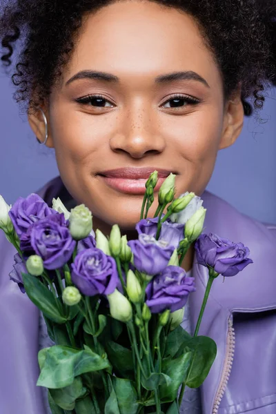 Усміхнена молода афроамериканка біля фіолетових квітів ізольована на фіолетовому — стокове фото