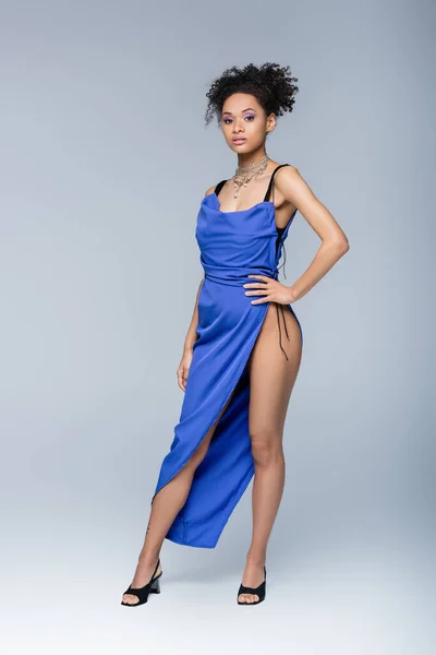 Comprimento total da mulher americana africana em vestido azul brilhante posando com a mão no quadril no cinza — Fotografia de Stock