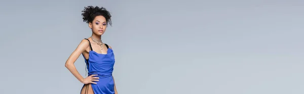 Elegante mujer afroamericana en vestido azul brillante posando con la mano en la cadera aislado en gris, bandera - foto de stock