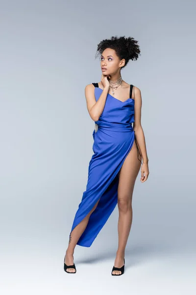 Longitud completa de mujer afroamericana joven tatuada en vestido azul posando en gris - foto de stock